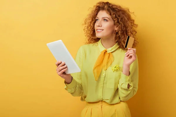 Dreamy redhead woman holding digital tablet and credit card on yellow — Stock Photo