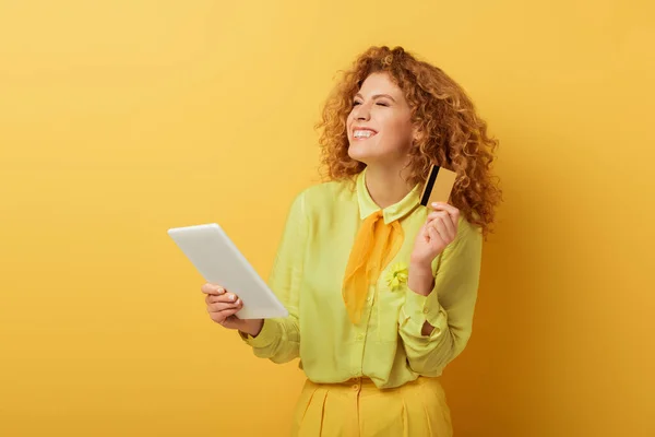 Улыбающаяся рыжая девушка держит цифровой планшет и кредитную карту на желтом — стоковое фото