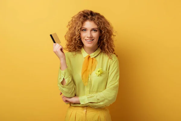 Sorridente ruiva menina segurando cartão de crédito no amarelo — Fotografia de Stock