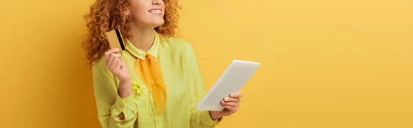 Plano panorámico de alegre pelirroja sosteniendo tableta digital y tarjeta de crédito en amarillo - foto de stock