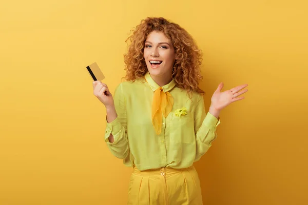 Excitada pelirroja sosteniendo tarjeta de crédito y gestos en amarillo - foto de stock