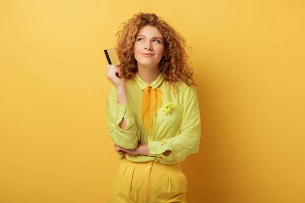 Sonriente pelirroja chica sosteniendo tarjeta de crédito mientras piensa en amarillo - foto de stock