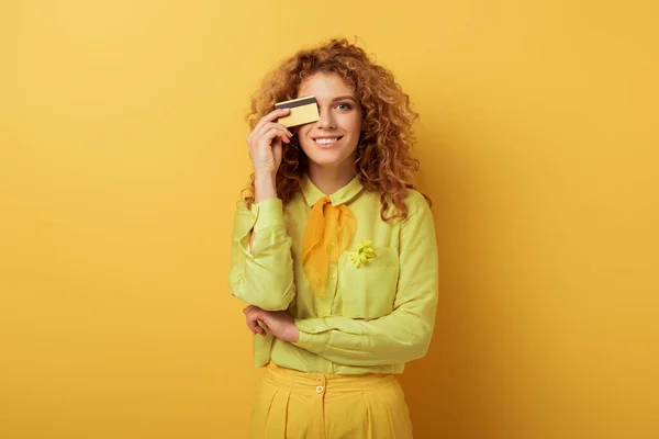 Lächelndes rothaariges Mädchen, das Augen mit Kreditkarte auf gelb bedeckt — Stockfoto