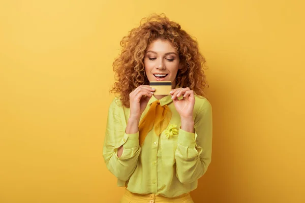 Positive redhead girl looking at credit card on yellow — Stock Photo
