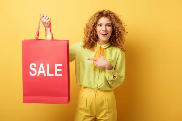 Cheerful redhead woman pointing with finger at shopping bag with sale lettering on yellow — Stock Photo