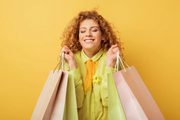 Happy redhead woman with closed eyes holding shopping bags isolated on yellow — Stock Photo