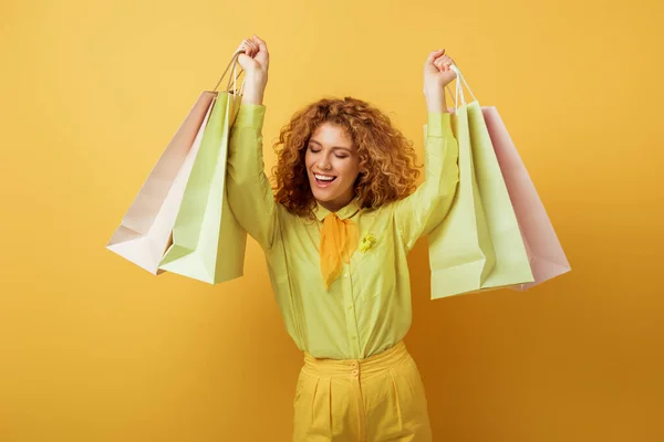 Счастливая рыжеволосая женщина с закрытыми глазами, держащая сумки с покупками на желтом — стоковое фото