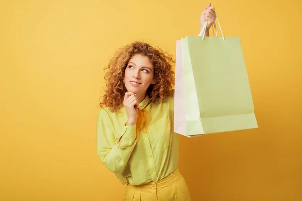 Задумчивая рыжая женщина держит сумки для покупок на желтом — стоковое фото