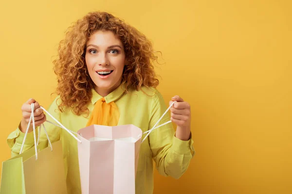 Aufgeregtes rothaariges Mädchen blickt in die Kamera und hält Einkaufstüten auf gelb — Stockfoto