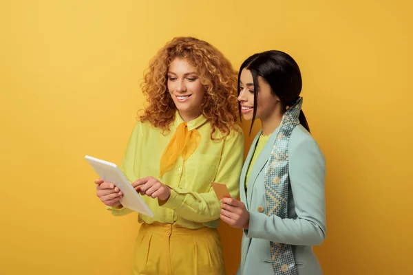 Heureux rousse fille en utilisant tablette numérique tout en faisant des achats en ligne avec afro-américaine détenant carte de crédit sur jaune — Photo de stock