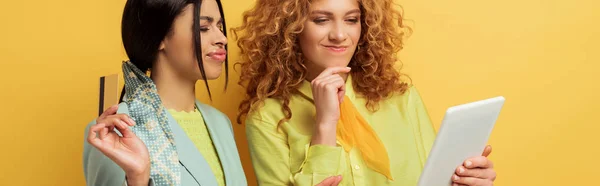 Colpo panoramico di ragazza rossa confusa utilizzando tablet digitale vicino attraente donna afro-americana in possesso di carta di credito isolata su giallo — Foto stock