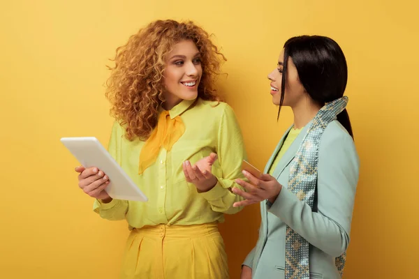 Happy redhead girl using digital tablet while looking at african american woman holding credit card on yellow — Stock Photo