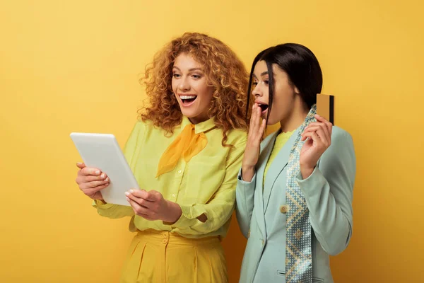 Alegre pelirroja chica usando digital tablet mientras que compras en línea con sorprendido africana americana mujer celebración de tarjeta de crédito en amarillo - foto de stock