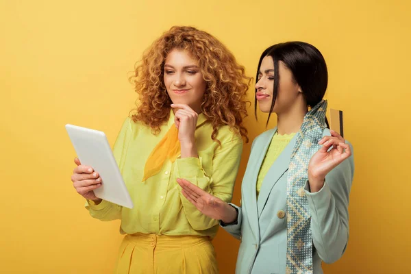 Confused redhead girl using digital tablet near attractive african american woman holding credit card on yellow — Stock Photo