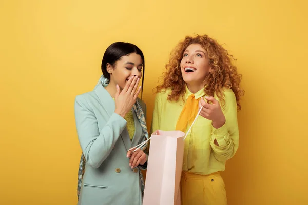 Cheerful redhead girl holding shopping bag near attractive african american woman laughing on yellow — Stock Photo