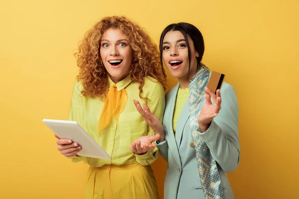 Surprised redhead girl using digital tablet near shocked attractive african american woman holding credit card on yellow — Stock Photo