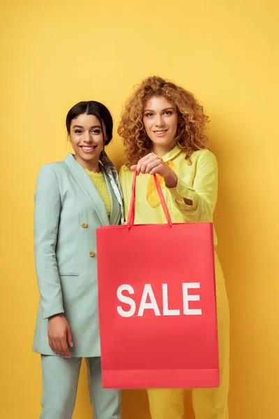 Menina ruiva feliz segurando saco de compras perto de sorrir mulher americana africana no amarelo — Fotografia de Stock