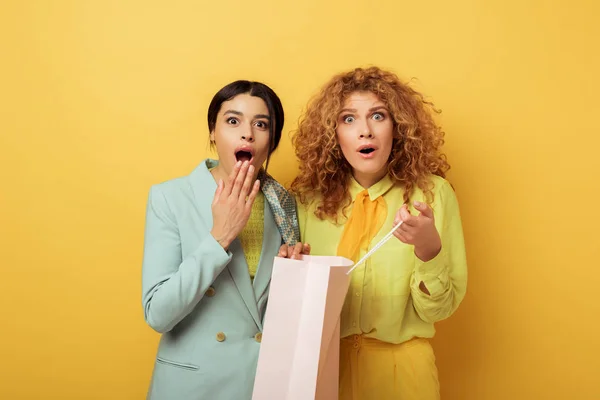 Shocked redhead girl holding shopping bag near surprised african american woman on yellow — Stock Photo