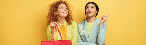 Disparo panorámico de chica pelirroja feliz sosteniendo bolsa de compras cerca de sonriente mujer afroamericana señalando con el dedo aislado en amarillo - foto de stock