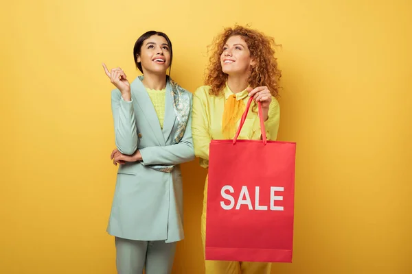 Chica pelirroja feliz sosteniendo bolsa de compras con letras venta cerca sonriente mujer afroamericana señalando con el dedo en amarillo - foto de stock