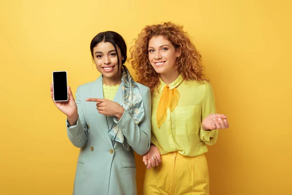 Glückliche afrikanisch-amerikanische Mädchen zeigt mit dem Finger auf Smartphone mit leerem Bildschirm in der Nähe Rotschopf Mädchen auf gelb — Stockfoto