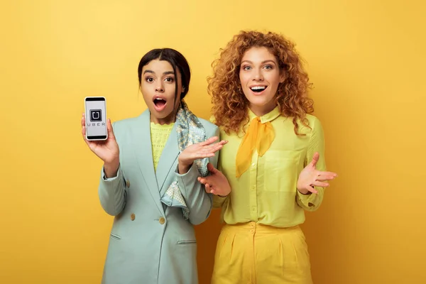 KYIV, UKRAINE - FEBRUARY 4, 2020: shocked african american girl holding smartphone with uber app on screen near surprised redhead girl on yellow — Stock Photo