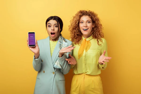KYIV, UKRAINE - FEBRUARY 4, 2020: shocked african american girl holding smartphone with instagram app on screen near surprised redhead girl on yellow — Stock Photo