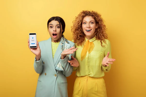 KYIV, UKRAINE - FEBRUARY 4, 2020: shocked african american girl holding smartphone with messenger app on screen near surprised redhead girl on yellow — Stock Photo