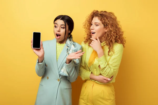 Impactado chica afroamericana mirando el teléfono inteligente con pantalla en blanco cerca de chica pelirroja sorprendida en amarillo - foto de stock