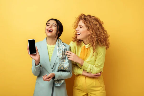 Happy african american girl holding smartphone blank screen and laughing with  redhead girl on yellow — Stock Photo