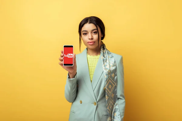 KYIV, UCRANIA - 4 de febrero de 2020: hermosa niña afroamericana sosteniendo teléfono inteligente con aplicación de youtube en amarillo - foto de stock