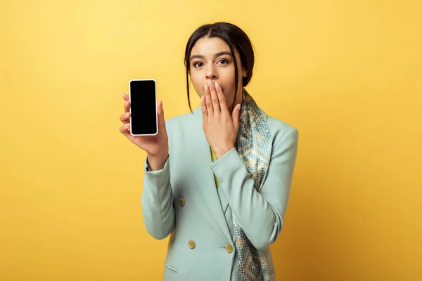 Hermosa chica afroamericana cubriendo la cara y sosteniendo el teléfono inteligente con pantalla en blanco en amarillo - foto de stock