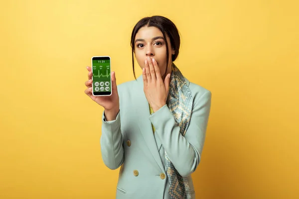 Chocado menina americana africana cobrindo rosto e segurando smartphone com aplicativo médico no amarelo — Fotografia de Stock