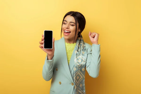 Chica afroamericana feliz sosteniendo teléfono inteligente con pantalla en blanco y celebrando en amarillo - foto de stock