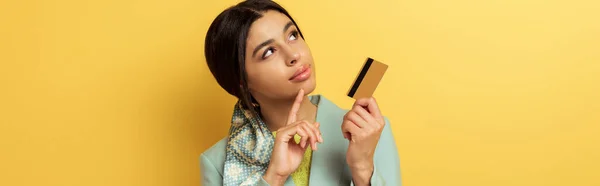 Panoramic shot of pensive african american girl holding credit card and pointing with finger on yellow — Stock Photo