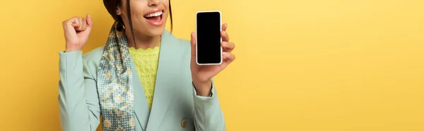 Plano panorámico de chica afroamericana feliz sosteniendo teléfono inteligente con pantalla en blanco y celebrando aislado en amarillo - foto de stock
