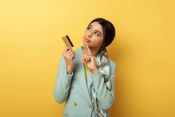 Chica afroamericana pensativa sosteniendo tarjeta de crédito y señalando con el dedo en amarillo - foto de stock