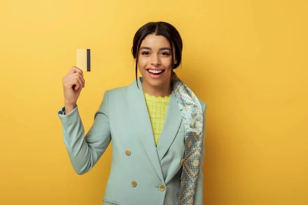 Chica afroamericana bonita sosteniendo tarjeta de crédito y sonriendo en amarillo - foto de stock