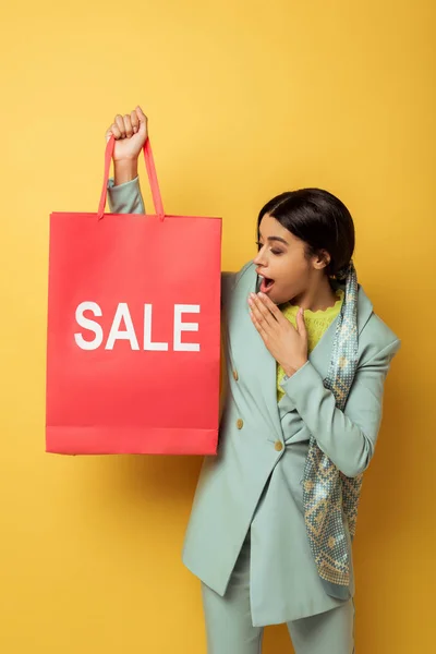 Surprised african american girl looking at shopping bag with sale lettering on yellow — Stock Photo