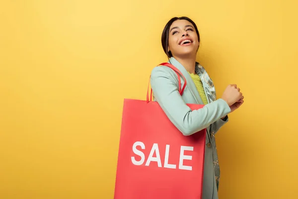 Cheerful african american girl holding shopping bag with sale lettering and smiling on yellow — Stock Photo