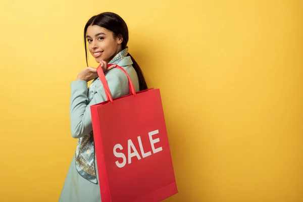 Positive african american girl holding shopping bag with sale lettering and smiling on yellow — Stock Photo