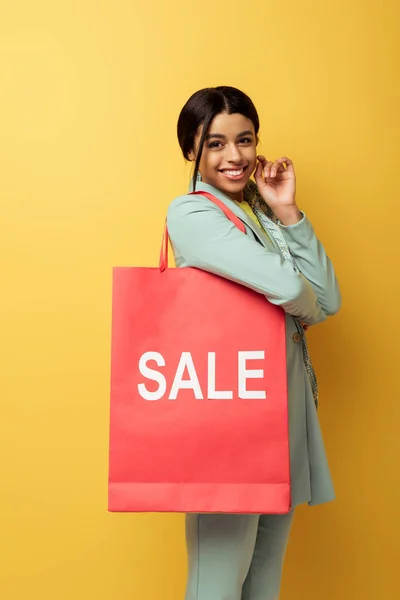 Positive african american woman holding shopping bag with sale lettering and smiling on yellow — Stock Photo