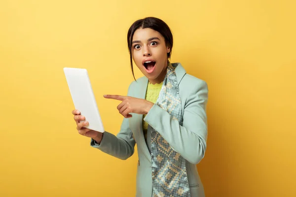 Mujer afroamericana sorprendida señalando con el dedo a la tableta digital en amarillo - foto de stock