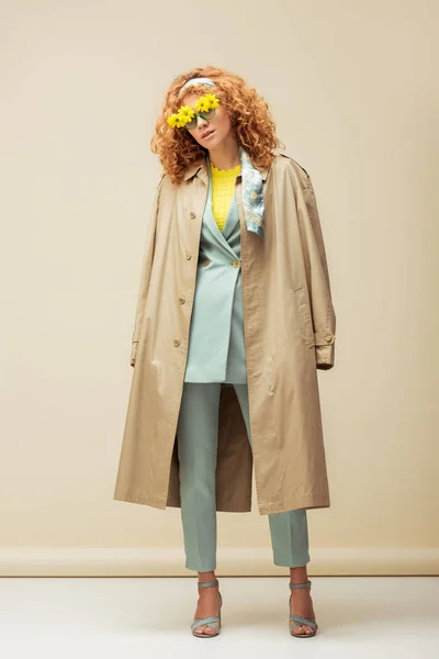 Stylisches rothaariges Mädchen in Sonnenbrille mit Blumen und Trenchcoat posiert auf beige — Stockfoto