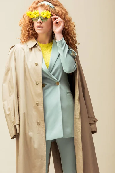 Élégant rousse femme en trench coat toucher des lunettes de soleil avec des fleurs tout en posant isolé sur beige — Photo de stock