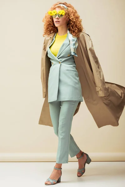 Stylish redhead woman in sunglasses with flowers and trench coat walking on beige — Stock Photo