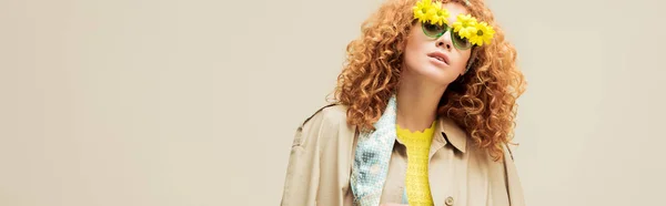Plano panorámico de mujer pelirroja con estilo en gabardina y gafas de sol con flores posando aisladas en beige - foto de stock