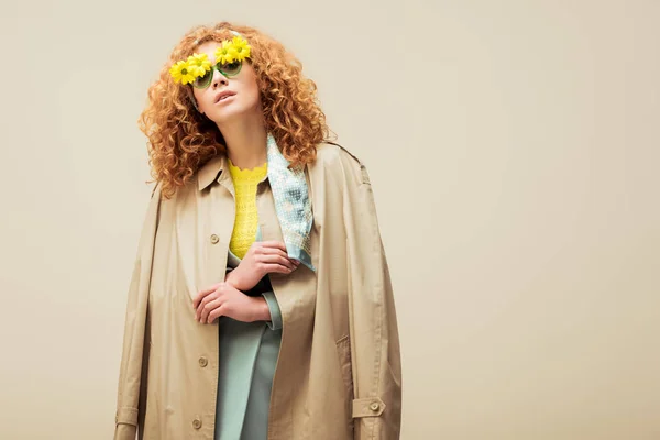 Stylische rothaarige Frau in Trenchcoat und Sonnenbrille mit Blumen posiert isoliert auf beige — Stockfoto