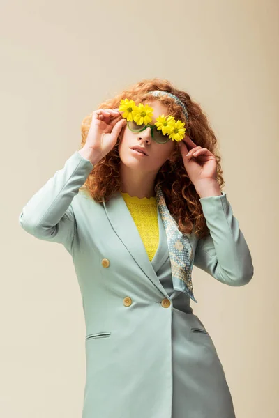 Stylish redhead woman in suit touching sunglasses with flowers while posing isolated on beige — Stock Photo