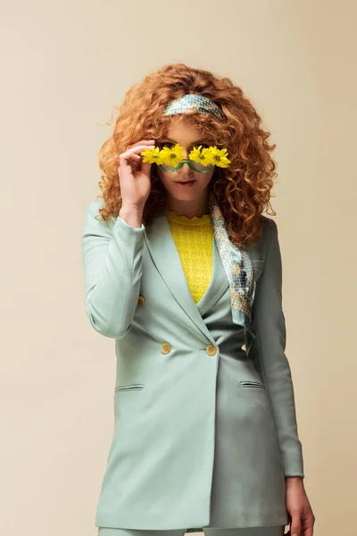 Mujer rizada y pelirroja en traje tocando gafas de sol con flores mientras posa aislada en beige - foto de stock